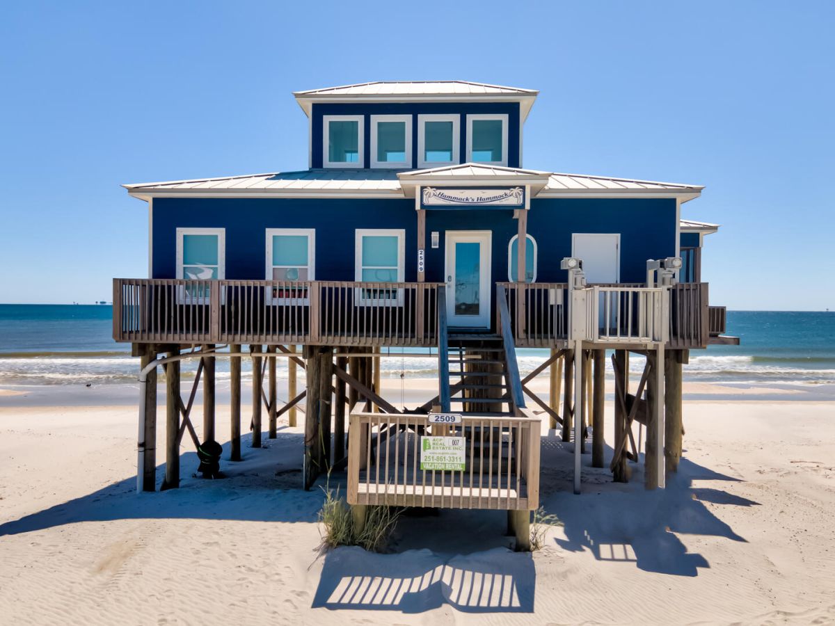 Blue beach house on stilts with a wooden deck and stairs leading to the sandy shore, ocean horizon in the background on a clear day.